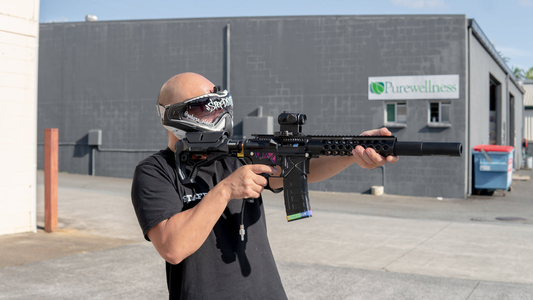 Person wearing a protective gel ball mask aiming a custom black gel blaster outdoors in an industrial area, standing in front of a building 